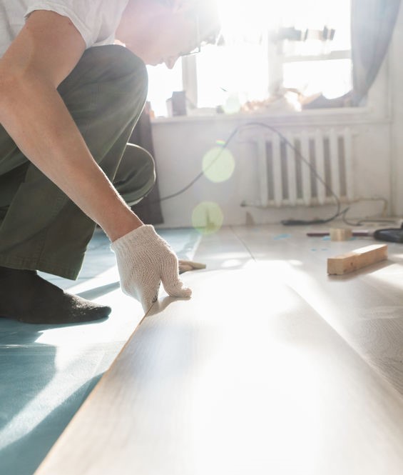 A person wearing work gloves installs a piece of light-hued wood flooring in a bright room.