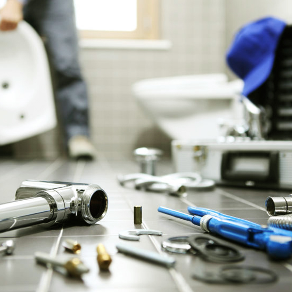 A plumber's tools and parts are spread on a bathroom floor. The background shows a person working near a sink.