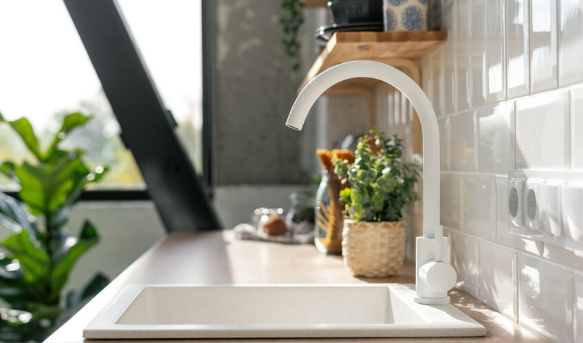 Side view of white sink in contemporary white kitchen interior. Wooden shelves with utensils. Spacious and bright apartment with plants concept A modern kitchen sink with a white faucet is set against a tiled backsplash. Wooden shelves hold various items and a plant is visible through the window.