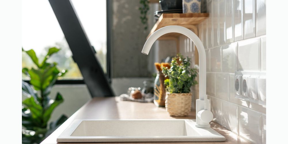 Side view of white sink in contemporary white kitchen interior. Wooden shelves with utensils. Spacious and bright apartment with plants concept A modern kitchen sink with a white faucet is set against a tiled backsplash. Wooden shelves hold various items and a plant is visible through the window.